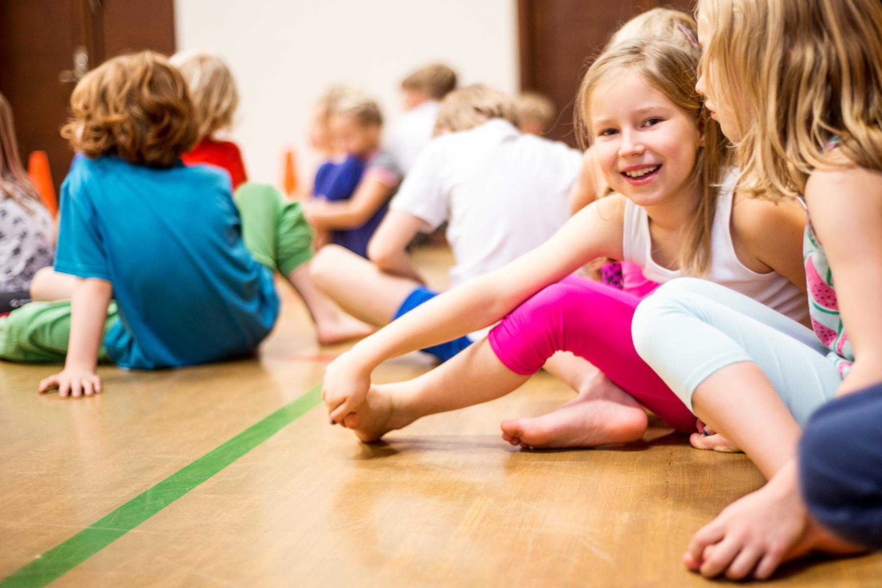 Lapset istuvat liikuntasalin lattialla ja tyttö katsoo hymyillen kameraan.
